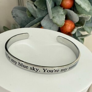 May include: A silver-colored cuff bracelet with the engraved text "You're my blue sky. You're my sunny". The bracelet is displayed on a white surface, with greenery and orange berries in the background.