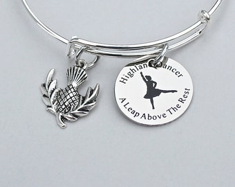 May include: Silver bangle bracelet with a charm featuring a stylized thistle and another charm with a silhouette of a Highland dancer and the text "Highland Dancer A Leap Above The Rest".