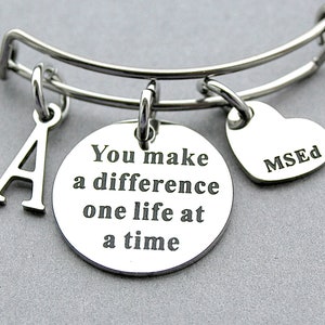 May include: A silver expandable bracelet with three charms: a letter A, a round charm with the text "You make a difference one life at a time", and a heart charm with the text "MSED".