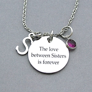 May include: A silver necklace with a charm that says "The love between Sisters is forever". The charm is attached to a silver chain with a small silver letter "S" and a small pink gemstone.