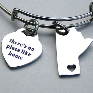 May include: A silver-toned bangle bracelet with two charms. One charm is a heart with the engraved text "there's no place like home." The other charm is a state outline with a heart cutout. The bracelet is on a gray background.