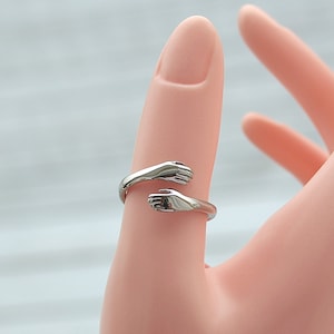 May include: Silver ring with two hands forming a hug, the hands are facing each other and the ring is open to fit on a finger.