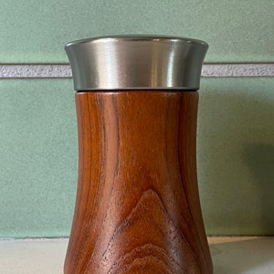 Może przedstawiać: Drewniany pojemnik na sól lub pieprz z srebrną pokrywką. Pojemnik ma zaokrągloną, rozszerzającą się podstawę i gładkie, polerowane wykończenie.