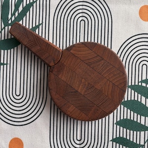 Puede incluir: Una tabla de cortar redonda de madera con un asa. La tabla es de madera marrón oscuro con un patrón de espiga. El asa es rectangular y está unida al lateral. La tabla está sobre un fondo blanco con un patrón negro y naranja.