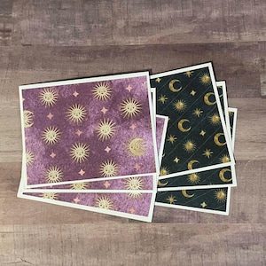 May include: A stack of square cards with celestial designs. One card features a purple background with gold suns, moons, and stars. Another card has a black background with gold moons, stars, and diamond patterns. The cards have white borders.
