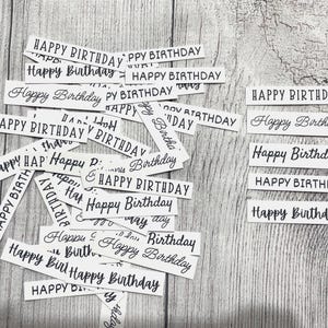 May include: A collection of rectangular white paper strips featuring the text Happy Birthday printed in various black fonts. The strips are arranged in a scattered pile on the left and a neat vertical stack of five on the right side.