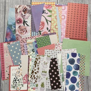 May include: A collection of rectangular paper strips featuring diverse patterns including floral prints, polka dots, geometric shapes, watercolor circles, and botanical illustrations of potted plants, with some pieces displaying handwritten script text.