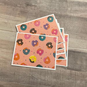 May include: A stack of square cards with a pink background and a repeating pattern of colorful donuts. The donuts have various colors, including pink, brown, teal, and yellow, with sprinkles. Each card has a white border.