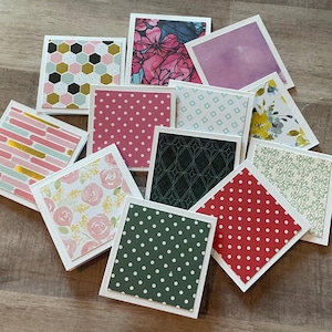 May include: A collection of square paper coasters with various patterns. Designs include floral, polka dots, geometric shapes, and abstract brushstrokes. The coasters have white borders and are arranged on a light-colored surface.