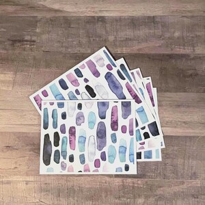 May include: A stack of watercolor art cards featuring an abstract design. The cards display a pattern of vertical brushstrokes in shades of blue, purple, pink, and black on a white background. The cards are arranged on a wooden surface.