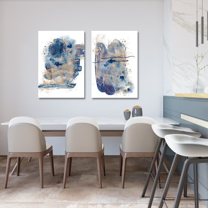 May include: Two abstract art prints featuring blue and gold watercolour paint splatters on a white background. The prints are framed and hanging on a white wall in a dining room.
