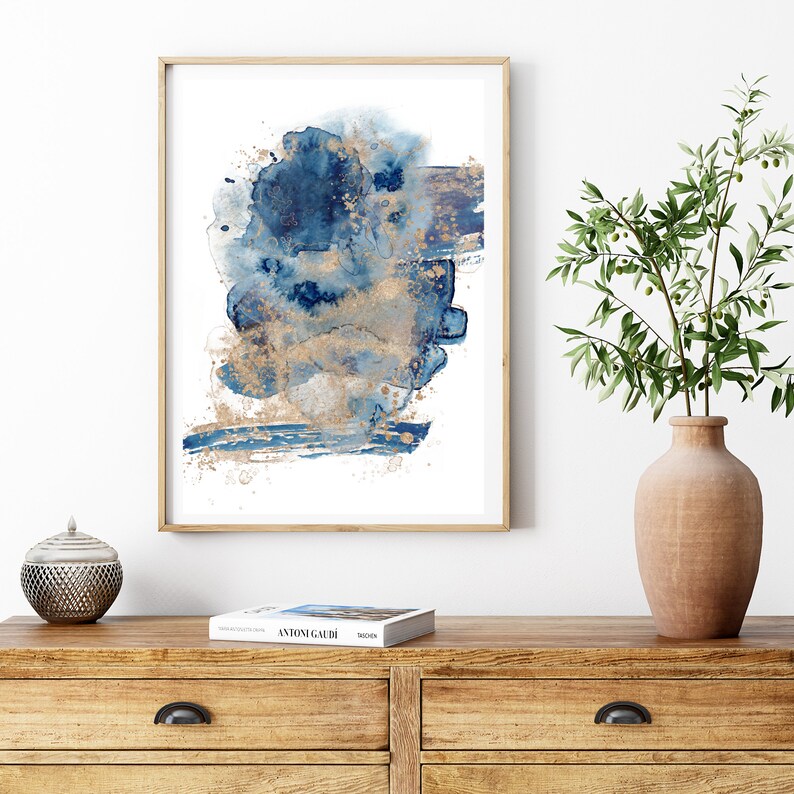 May include: Abstract watercolour painting in shades of blue and gold. The painting features a large, abstract shape in the centre, with smaller shapes and details surrounding it. The painting is framed in a simple, wooden frame.