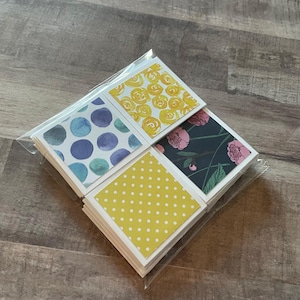 May include: A stack of square cards with various designs, including yellow roses, blue watercolor circles, pink floral patterns, and yellow polka dots. The cards are packaged in a clear plastic sleeve.