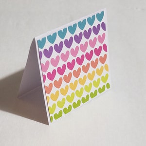 Op de afbeelding: Een witte kaart met een regenboogpatroon van harten. De harten zijn in tinten blauw, paars, roze, oranje, geel en groen.