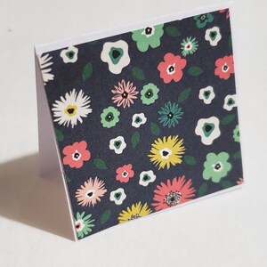Op de afbeelding: Een donkerblauwe kaart met een witte rand heeft een herhalend patroon van kleurrijke bloemen en bladeren. De bloemen zijn roze, geel, groen en wit.