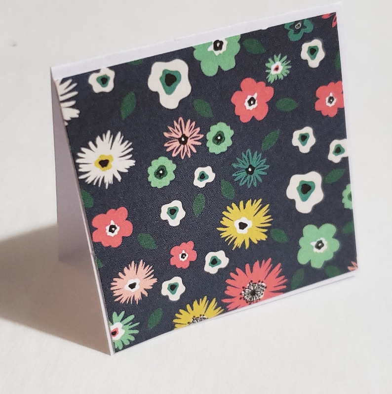 Op de afbeelding: Een donkerblauwe kaart met een witte rand heeft een herhalend patroon van kleurrijke bloemen en bladeren. De bloemen zijn roze, geel, groen en wit.