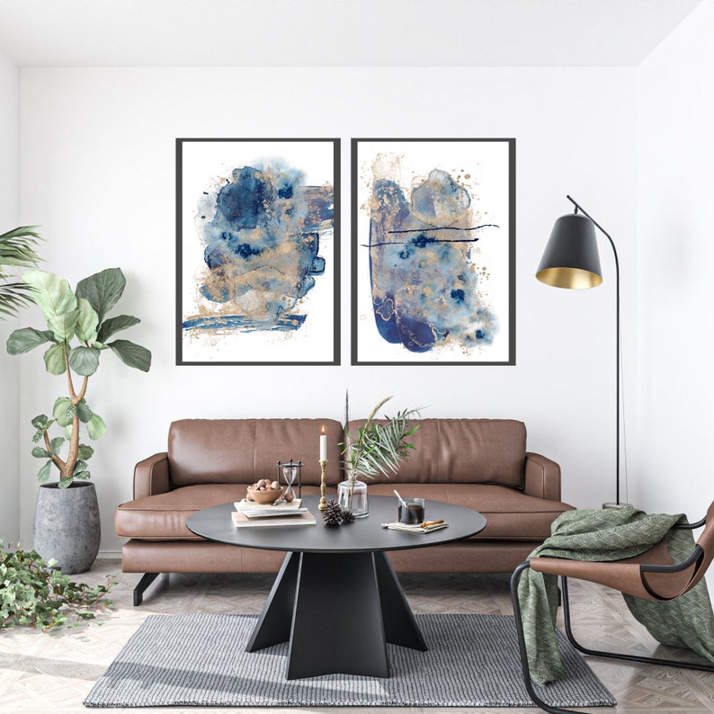 May include: Two abstract art prints featuring blue and gold watercolour designs. The prints are framed in black and hung on a white wall above a brown leather sofa.