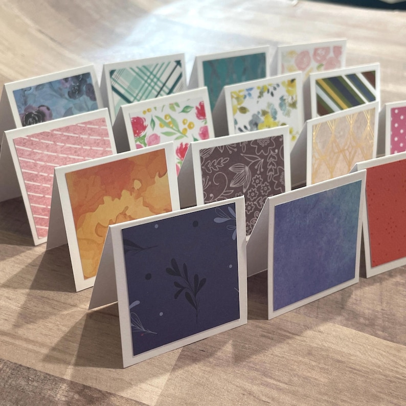 May include: A collection of small, folded greeting cards with various patterned designs. The cards feature floral, geometric, and abstract patterns in colors like blue, pink, orange, and green. The cards are arranged on a wooden surface.