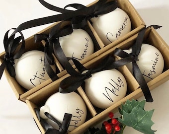 May include: A set of six white Christmas ornaments, each with a name in black script, presented in a divided cardboard box. Black satin ribbons are tied in bows around each ornament. A sprig of holly with red berries adds a festive touch.