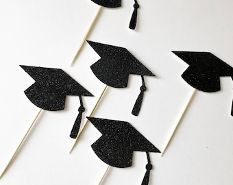 May include: Five black graduation cap toppers with gold glitter. The caps are attached to wooden skewers.