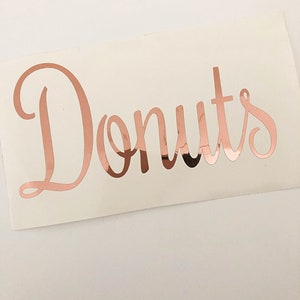 May include: A rose gold vinyl sticker with the word "Donuts" written in a cursive font.
