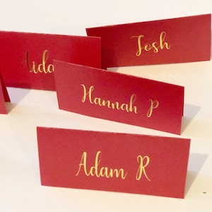May include: Four red place cards with gold lettering. The names on the cards are Ida, Josh, Hannah P, and Adam R.