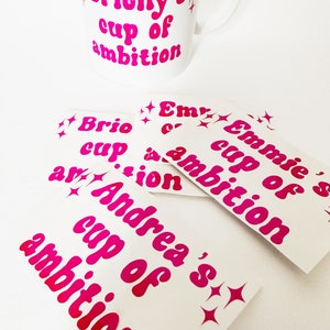 Puede incluir: Un conjunto de pegatinas de vinilo rosa con el texto "cup of ambition" y varios nombres. Las pegatinas están sobre un fondo blanco.