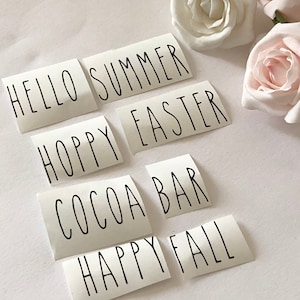 May include: Five white rectangular stickers with black text. The stickers read "Hello Summer", "Hoppy Easter", "Cocoa Bar", "Happy Fall", and "Happy Fall".