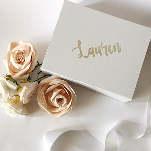 May include: A white gift box with the name "Lauren" written in gold lettering. The box is surrounded by pale peach silk flowers and a white satin ribbon.