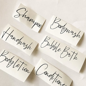 May include: Six white labels with black handwritten text for bathroom products. The labels read "Shampoo", "Bodywash", "Handwash", "Bubble Bath", "Bodylotion", and "Conditioner".