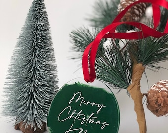 May include: A round, clear glass bauble with a green painted background and white lettering that reads "Merry Christmas Kelly". The bauble is hanging from a red ribbon.