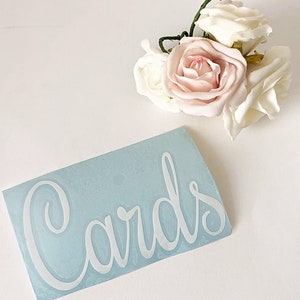 May include: A light blue rectangular sign with the word "Cards" in white cursive script. A cluster of artificial roses in shades of white and blush pink is positioned above the sign, with green stems visible.