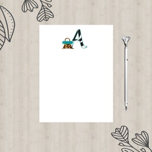 May include: A white notepad with a black and white patterned letter A and a small brown and black patterned bag on a wood background.