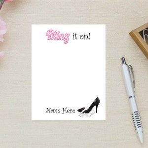 May include: A white card with the text "Bling it on!" in pink lettering. The card has a black high heel shoe with a pearl necklace on the bottom right corner. The text "Name Here" is written below the shoe.