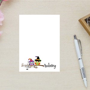 May include: A white notepad with a Halloween-themed illustration of two owls wearing witch hats and sitting on a branch. The text "Aubrey" is written below the owls.