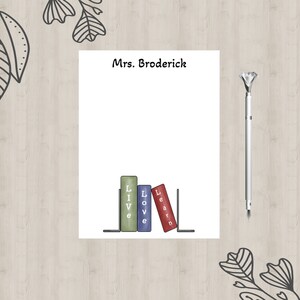 May include: A white notepad with the text "Mrs. Broderick" at the top. The notepad has a design of three books with the words "Live", "Love", and "Learn" on them. The books are on a black bookshelf.