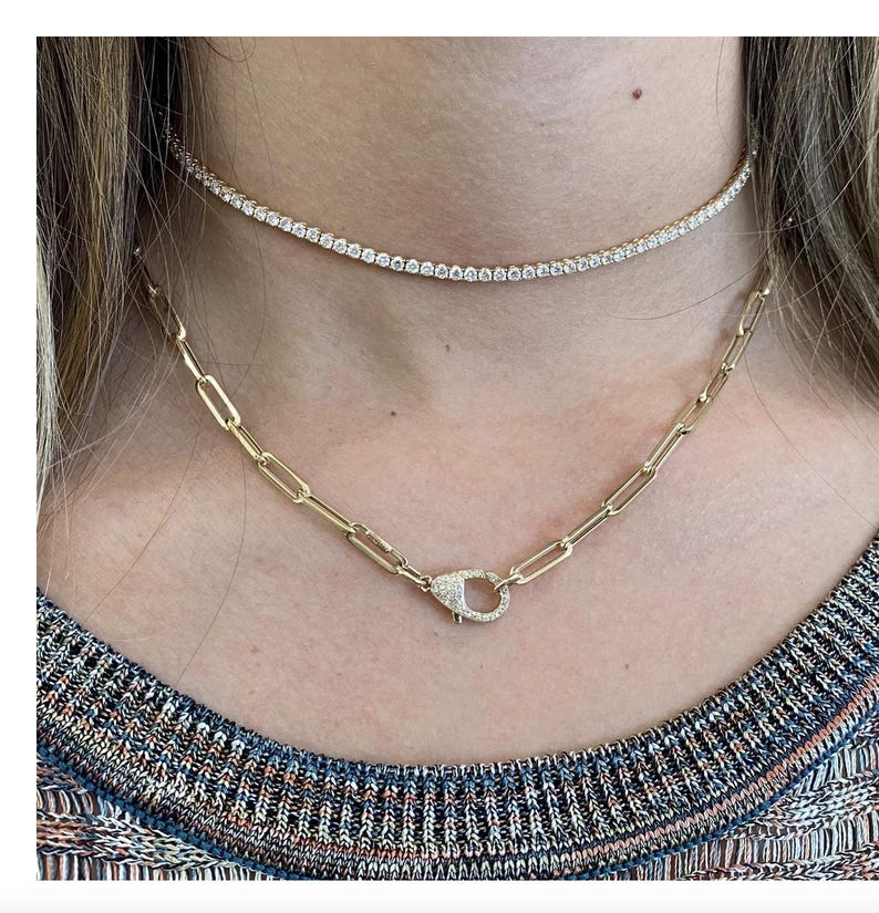 May include: Two gold necklaces with diamond accents. The top necklace is a thin, delicate chain with small diamonds. The bottom necklace is a thicker chain with a larger, diamond-encrusted clasp.