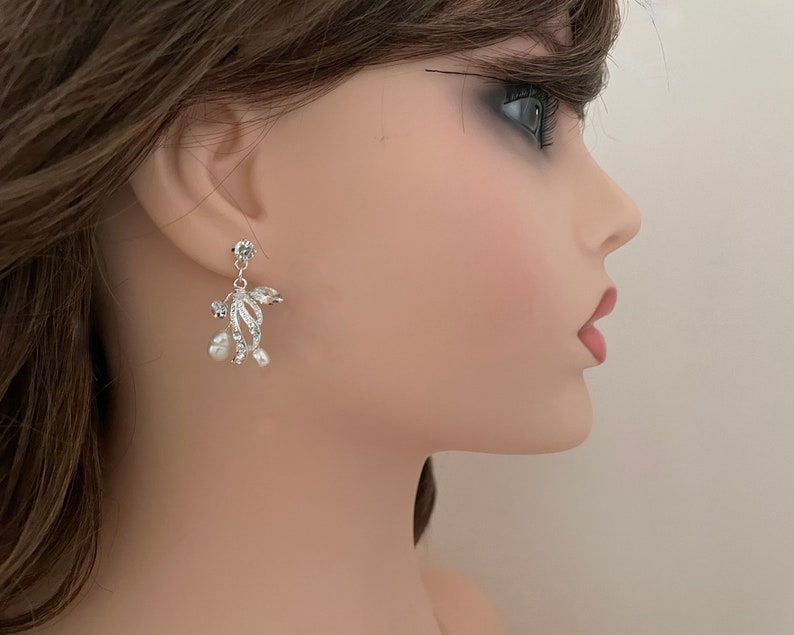 May include: Silver-toned dangle earrings with a floral design. The earrings feature clear crystals and small, white pearls. The earrings are attached to a small, round crystal stud.