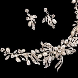 May include: A silver-toned bridal jewelry set featuring a necklace and matching earrings. The necklace and earrings are adorned with faux pearls and clear crystals, arranged in a floral and leaf design. The set is displayed against a black background.