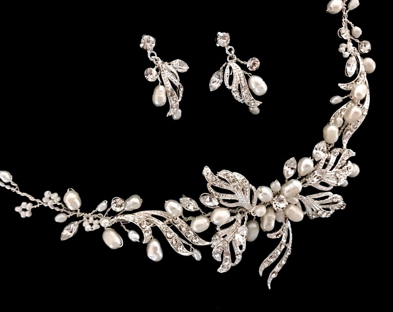 May include: A silver-toned bridal jewelry set featuring a necklace and matching earrings. The necklace and earrings are adorned with faux pearls and clear crystals, arranged in a floral and leaf design. The set is displayed against a black background.