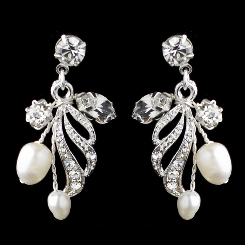 May include: Elegant silver-toned drop earrings with a floral design. Each earring features a large, round clear crystal at the top, with smaller crystals and a leaf-like design. Two white, teardrop-shaped pearls dangle from each earring. The background is black.