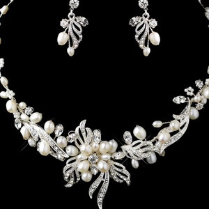 May include: A silver-toned necklace and earring set. The necklace features a floral design with white pearls and clear crystals. The earrings have a similar design with pearls and crystals. The jewelry is set against a black background.