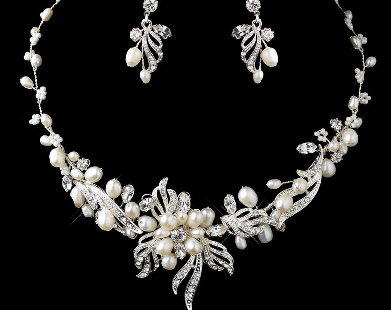 May include: A silver-toned necklace and earring set. The necklace features a floral design with white pearls and clear crystals. The earrings have a similar design with pearls and crystals. The jewelry is set against a black background.