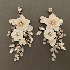 Puede incluir: Un par de pendientes de tono dorado con detalles de flores blancas y rosadas y cristales transparentes. Los pendientes son colgantes y tienen un diseño delicado y femenino.