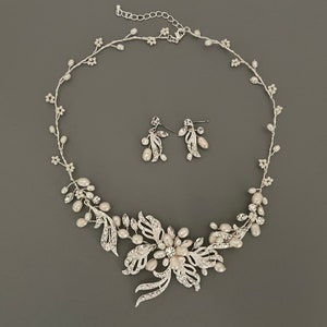 May include: A silver-toned bridal jewelry set featuring a necklace and matching earrings. The necklace has a floral design with faux pearls and clear crystals. The earrings have a leaf and pearl design. The set is displayed on a gray background.