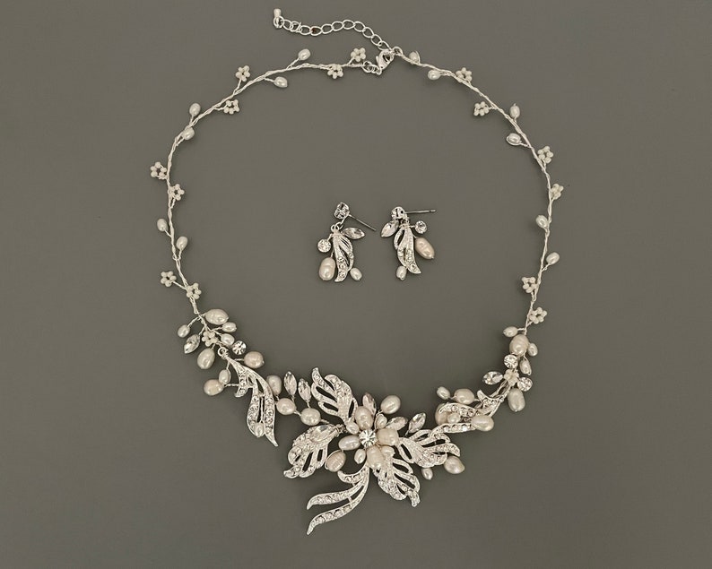 May include: A silver-toned bridal jewelry set featuring a necklace and matching earrings. The necklace has a floral design with faux pearls and clear crystals. The earrings have a leaf and pearl design. The set is displayed on a gray background.