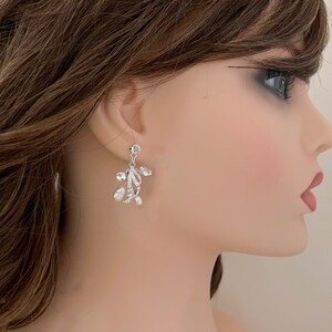 May include: Elegant silver-toned earrings with a floral design. Each earring features clear crystals and small, dangling pearls. The earrings have a delicate, sophisticated appearance, suitable for special occasions.