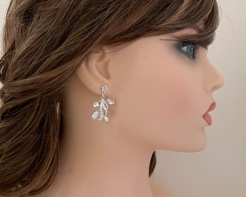 May include: Elegant silver-toned earrings with a floral design. Each earring features clear crystals and small, dangling pearls. The earrings have a delicate, sophisticated appearance, suitable for special occasions.