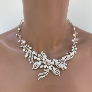 May include: A silver-toned necklace with a floral design. The necklace features clear crystals and white pearls. The central floral element is surrounded by delicate, vine-like strands with pearls. The necklace is designed to be worn around the neck.