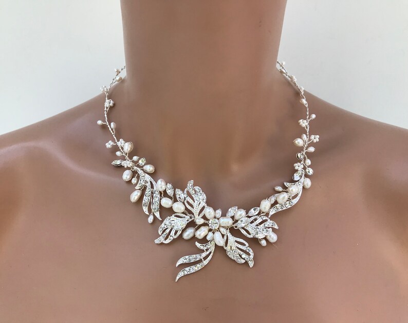 May include: A silver-toned necklace with a floral design. The necklace features clear crystals and white pearls. The central floral element is surrounded by delicate, vine-like strands with pearls. The necklace is designed to be worn around the neck.